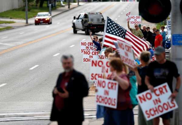 A group of white protesters stands by a road holding signs that read "No CRT in Schools"