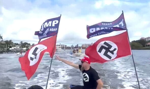 A Nazi in a Trump hat salutes from the back of a boat that is flying Trump flags and Nazi flags.